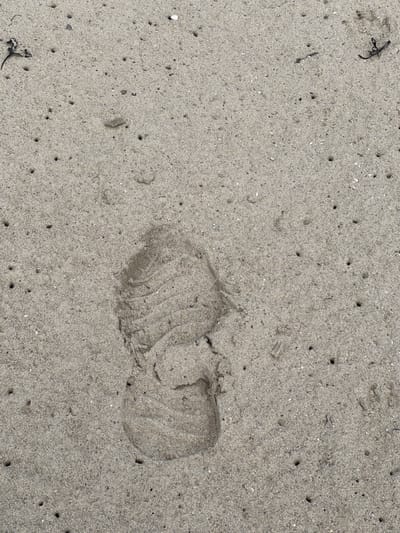 A footprint is imprinted in the sand of a beach in Tullycullion, Ireland. The footprint, likely from a shoe, is clearly visible and shows the tread pattern. The surrounding sand is dotted with small holes, suggesting the presence of marine life or erosion. Small pieces of debris, possibly seaweed or shells, are scattered on the sand. The scene is outdoors, with natural lighting.
