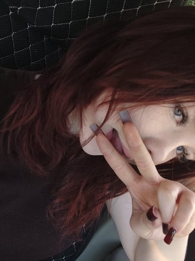 A close-up, tilted, chest-up shot shows a person with long, dark red hair and light skin. They are wearing a dark top and have long, dark red, square-shaped nails. Their right hand is held up to their face, with two fingers extended in a peace sign. The person's mouth is slightly open, with their tongue visible. Their eyes are light blue and appear to be looking to the right. The background consists of a dark, textured material with a white diamond-shaped pattern. There are no discernible landmarks or features to indicate a specific location in Shimanovsk, Russia.