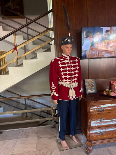 The image displays an indoor scene in Melnik, Bulgaria, featuring a full-body mannequin dressed in historical military attire positioned on a light-colored tiled floor. The mannequin wears a long-sleeved red jacket adorned with multiple rows of white cording and buttons, dark blue trousers with a white side stripe, and a white braided sash with tassels. Its head is covered by a tall grey fur hat featuring a large dark feather, a red, white, and green tricolor ribbon, and a metallic star-shaped emblem. The mannequin's bare feet are visible, resting on a clear base.

To the left of the mannequin, a multi-level staircase with white steps and railings made of dark and golden-colored metal bars ascends. Several red ribbons are tied to the railing bars at different heights. A framed painting of a historical figure is partially visible on the wall above the upper section of the stairs.

To the right of the mannequin, a dark wooden paneled wall serves as a backdrop. Mounted on this wall is a flat-screen television (JVC brand visible) displaying an image of three individuals (two adults and one child) gathered around a table set with food and drinks. Below the television, a polished wooden chest of drawers with intricate inlays and ornate brass handles is present. On its top surface, two books are visible; one is titled "TERVEL THE SAVIOR OF MEDIEVAL EUROPE," and another, partially obscured, shows "BULGARIAN" in its title. Adjacent to the books is a decorative tray containing small gourds, flowers, and candles.
