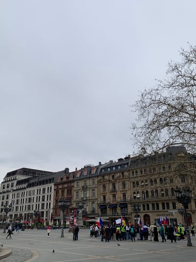 The image shows a group of people gathered in a large open plaza in Frankfurt am Main, Germany, located at 50.115 latitude and 8.671 longitude. The plaza is surrounded by tall buildings with ornate facades. The majority of the people are standing in a group in the center of the plaza, many holding flags. The sky is overcast with clouds. The building on the left is a white office building, while the buildings in the middle and right are yellow and orange with ornate designs and large windows. There are trees without leaves visible in the background. There is a small pigeon in the foreground. The plaza appears empty, with no traffic or other people.