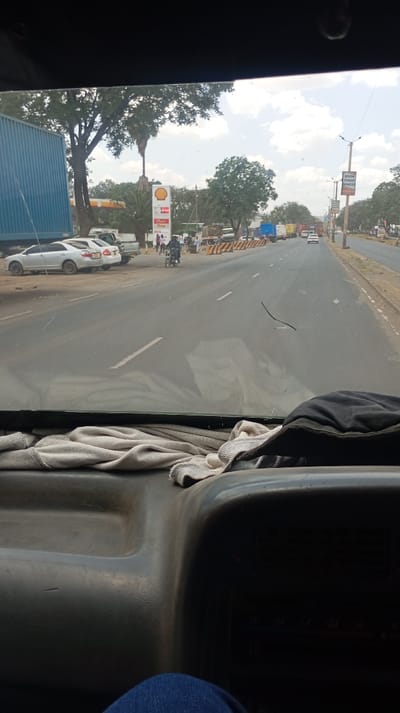A view from inside a vehicle shows a straight asphalt road with cars and trucks lined up on the left side. A Shell gas station is visible with its distinctive logo. Trees line both sides of the road, adding to the outdoor setting. The foreground features a partially visible dashboard with a cloth and a glimpse of the driver's leg. The overall scene suggests a traffic jam near a gas station on a sunny day.