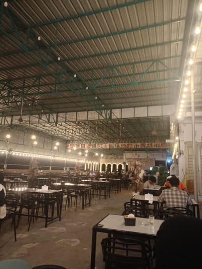 The image displays a large, high-ceilinged dining area within a restaurant in Namplohbaro, Indonesia, observed at night. The ceiling features exposed green metal trusses supporting a corrugated metal roof, with numerous white globe pendant lights and strings of decorative incandescent bulbs providing illumination. The floor is concrete, showing irregular lighter markings. The space is furnished with multiple rows of dark-framed tables with light-colored tops and accompanying dark chairs. Several individuals are present, primarily seated at tables in the mid-ground and background, engaged in dining or conversation. Specifically, in the mid-ground right, a person in a plaid shirt is seated with their back to the viewer, alongside another individual in a dark shirt looking downwards, and a third person partially obscured, wearing an orange top and a cap. On various tables, tissue boxes and clear glasses are visible. The background reveals an architectural feature composed of several illuminated arches. Above these arches, a series of approximately 15 framed items, likely menus or pictures, are displayed horizontally. To the far right, a vertical pillar features a sign reading "KASIR" (Cashier). Decorative potted plants or artificial trees are strategically placed throughout the dining area.