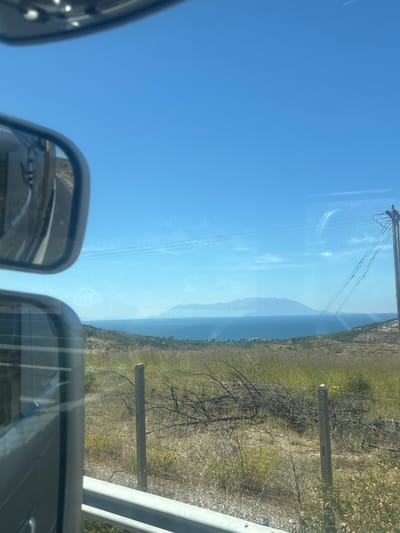 The image, taken from a vehicle, captures a scenic view of the sea and a distant mountainous coastline under a clear blue sky. In the foreground, a side-view mirror is visible on the left, reflecting the sky and the interior of the vehicle. Below it, another mirror or window reflects a similar view. The landscape consists of dry, grassy hills with scattered bushes and some dead branches. Two concrete posts with wires are visible in the middle ground, part of a fence or utility line. In the distance, the calm, blue sea stretches out, meeting a hazy, mountainous landmass. A few buildings are discernible on the coastline. The overall impression is one of a sunny, rural coastal environment. While the image is identified as being taken in Avra, Greece, there are no specific landmarks or details within the image that definitively confirm this location. The scene is devoid of any people.