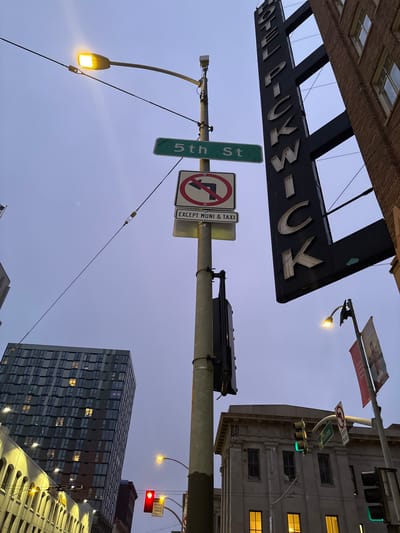 This is a low-angle, wide shot of a city street scene in San Francisco, USA. The sky is overcast and appears to be dusk. In the foreground, a lamppost holds multiple street signs. A green sign reads "5th St". Below it is a circular sign with a red border and a red diagonal line over a black arrow pointing left, indicating no left turn. Underneath this is a white rectangular sign reading "EXCEPT MUNI & TAXI".
To the right of the lamppost, a tall, black vertical sign with large white letters reads "HOTEL PICKWICK". The sign is illuminated by internal lighting, though some letters are damaged or not lit. Attached to the sign are several electrical wires. A brick building is visible to the right of the sign.
In the background, a modern, tall glass building with many windows, some lit, is visible on the left. Below and to the right of the Hotel Pickwick sign, a street light is illuminated with a yellow bulb. Further to the right, another lamppost displays a red banner with an image of the Golden Gate Bridge. A traffic light with a green light illuminated is visible in front of a classical-style building. Another traffic light with a red signal is visible further to the right. The overall scene depicts an urban environment with various commercial establishments and traffic control devices.