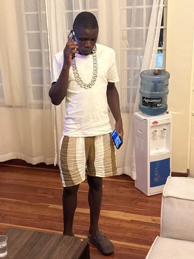 A man stands in a room with a white curtained window behind him. He is holding a phone to his ear with his right hand and looking at another phone in his left hand. He is wearing a white t-shirt, a large silver chain, and striped shorts. A water dispenser sits against the window, and a couch is in the background. The floor is wooden.