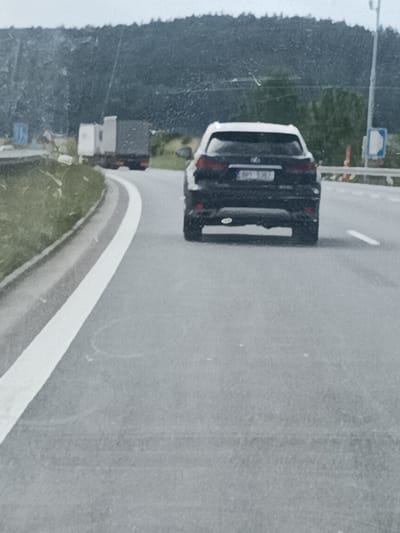 This media file captures a rear view of a black Lexus SUV driving on a paved road. The SUV is positioned in the right lane, moving away from the camera. Its license plate is visible and reads "BP1 1347". In the background, a large truck with a light-colored cab and a dark cargo area is also visible, driving ahead and to the left of the SUV. The road curves to the left, bordered by a grassy embankment on the left and a barrier with traffic signs on the right. The sky appears overcast, suggesting a cloudy or possibly rainy day. The overall scene depicts a moment of road travel, likely on a highway or a major road, with a forested hill in the far background. The lighting is diffused, typical of an overcast sky. The image quality is somewhat grainy, suggesting it was taken with a mobile device or under low-light conditions. There are no people clearly visible in the image.