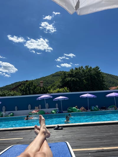 The image depicts a bright, sunny day at a poolside location in Varvara, Bulgaria. In the foreground, a person's legs and feet are visible, relaxed on a blue lounge chair. The pool, filled with clear blue water, is the central focus, with several people enjoying themselves. Some are swimming, while others are lounging on beanbag chairs around the pool's edge. Purple umbrellas, some with "Ice Cream" branding, provide shade. A vibrant blue wall lines the pool area, separating it from the surrounding greenery and a hill in the background. A house is partially visible amidst the trees on the hill. The sky is a brilliant blue with scattered white clouds. The overall scene conveys a sense of leisure and summer enjoyment.