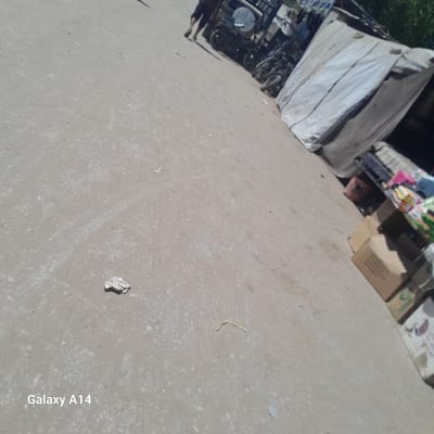 The image depicts an outdoor scene in what appears to be a market or a street with stalls. The primary subjects are various objects, including what looks like a stall covered with a large white tarp, possibly sheltering goods. Behind this, other stalls or structures with more items, including cardboard boxes and what seem to be products like cleaning supplies or toys, are visible. In the foreground and mid-ground, a grey, dusty ground surface is prominent, with scattered debris and small pebbles.

To the left of the frame, a person is partially visible, with their legs and feet wearing dark pants and shoes, suggesting they are standing or walking on the ground. Near this person, several bicycles or motorcycle wheels are visible, suggesting some form of transport or personal belongings are present.

The overall setting is somewhat disorganized, typical of a marketplace, with a mix of covered and open areas. The ground is uneven and appears to be a dirt or unpaved surface. The lighting suggests it is daytime, with sunlight creating some shadows, although the overall brightness indicates a clear or partly cloudy sky. There is no strong indication of specific weather conditions beyond the dusty ground.

In the lower right corner of the image, white text is present, reading "Galaxy A14". This appears to be a watermark or text added by the device that captured the image, indicating it was taken with a Samsung Galaxy A14. There are no other discernible text elements within the scene itself, apart from what might be on the packaging of the goods in the stalls, which is too blurry to read. The image conveys a sense of everyday commerce and activity in an informal setting.