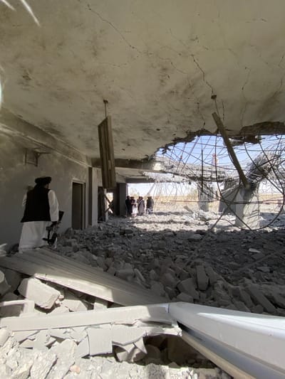 The scene depicts the interior of a severely damaged concrete building in Mandisar, Afghanistan. The floor is covered by a deep layer of shattered concrete and construction debris. The ceiling is extensively cracked, with a significant section having collapsed, revealing twisted rebar and the clear sky. A rectangular wooden panel hangs vertically from a damaged point in the ceiling. In the left foreground, an individual wearing a white shalwar kameez, a dark vest, and a dark turban stands amidst the rubble, holding a rifle with a visible scope. This individual is positioned facing right. In the mid-ground, a group of approximately five to six individuals, similarly dressed in traditional clothing, are standing near a brightly lit opening further within the damaged structure. Beyond the collapsed area and openings, remnants of concrete pillars and a dry, arid outdoor environment are visible.