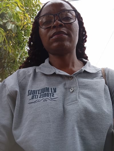 A medium shot captures a Black woman from the chest up, looking directly at the camera. She wears glasses with thick black frames and her dark, braided hair cascades down her shoulders. Her skin appears dewy, suggesting sweat or moisture, possibly from the heat. She is wearing a light gray polo shirt with the collar visible. Embroidered on the shirt, in a dark, possibly navy blue, font, is the text "VI MULTIJUM I.V." above what appears to be "OTI 2380T2", with decorative swirls beneath. The background is a blur of green foliage from trees on the left, and a bright, overcast sky on the right. The scene suggests an outdoor setting, likely during the day, given the natural light. The woman's expression is neutral, with her lips slightly parted.