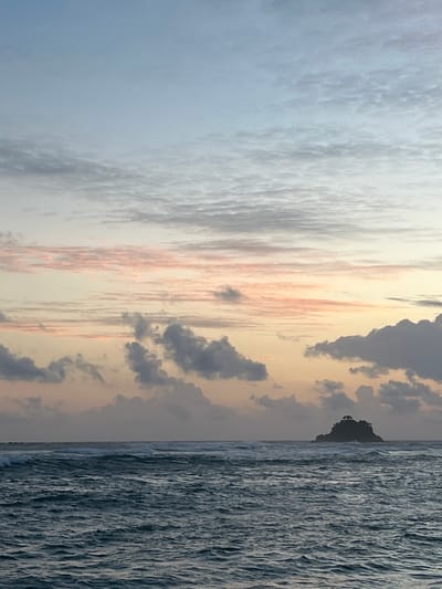 This media file captures a serene coastal scene at what appears to be either sunrise or sunset, given the soft, warm hues in the sky. The image is oriented vertically.

The scene is divided into two distinct parts. The left side of the frame is dominated by a vast expanse of sky filled with textured clouds. These clouds range in color from a soft, pale blue and white on the far left to warmer tones of peach and pale yellow further to the right, suggesting the sun's rays are interacting with them. Darker, more ominous clouds are also present, particularly on the right side of the sky.

The right side of the frame is entirely occupied by the ocean. The water is depicted with a great deal of detail, showing the texture of choppy waves with a silvery-blue sheen, indicating the presence of light reflecting off the surface. The line where the sky meets the ocean is visible, and in the distance, a small, dark silhouette of what appears to be a rocky outcrop or small island breaks the horizon.

The overall mood is one of tranquility and natural beauty. The time of day is suggested by the lighting and cloud colors. The location is identified as Midigama, Sri Lanka, which is a coastal area, consistent with the imagery of the ocean and sky. There is no visible text in the media file. The focus is purely on the natural landscape, with no people or man-made objects prominently featured, other than the distant island.