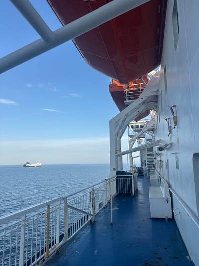 This is a photograph taken from the deck of a ferry, looking out towards the sea. The perspective is from a side-facing angle, with the bulk of the ferry's superstructure dominating the right side of the frame. The deck is painted a deep blue and appears wet in places, with a white metal railing running along the edge.

A large, orange life raft is suspended above the deck on a white metal davit system. Further along the deck, there are white benches and various pieces of equipment, including what appears to be a fire extinguisher. The side of the ferry is painted white, with several rectangular windows visible.

In the distance, on the calm, blue sea, another ship is sailing. The sky above is a clear, bright blue with a few wispy white clouds. The scene suggests a journey across the water, possibly in a coastal region of Sweden, given the presence of ferries in this area. The overall impression is one of open water and maritime travel.