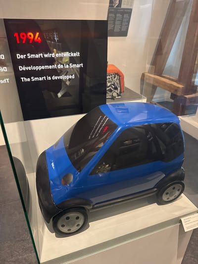 This media file depicts a blue model of a Smart car displayed in a museum exhibit. The car is positioned on its side, showcasing its sleek design and black tinted windows. In the background, wooden beams are visible, suggesting an older architectural setting.
A prominent display panel to the left of the car features text in three languages: German, French, and English, all stating "Der Smart wird entwickelt," "Développement de la Smart," and "The Smart is developed," respectively. Above these phrases, the number "1994" is displayed in large red font, indicating the year associated with the Smart car's development. Further text on the display panel, partially obscured, appears to provide historical context related to the car's model and its manufacturing origins in Grenchen, Switzerland, with a reference to the "Berna Historical Museum."
The scene is well-lit, likely within an indoor museum environment. There are no people visible in the immediate frame, focusing the attention entirely on the exhibit. The overall impression is one of historical preservation and presentation of an iconic automotive design.