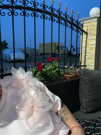 The image shows a close-up of a person wearing a white garment adorned with large, sheer pink fabric flowers, one of which is in focus in the foreground. The person's arm, with a tattoo of flowers on it, is visible to the right. They are wearing a red string bracelet. The scene appears to be outdoors, possibly on a patio or balcony, with a black wrought-iron fence in the foreground. Beyond the fence, a building with a stone facade and a round light fixture is visible against a clear blue sky. In the background, there are trucks and what looks like a potted plant with red flowers. The overall impression is that of a casual outdoor setting, perhaps in the late afternoon or early evening, given the lighting. The image is oriented vertically.