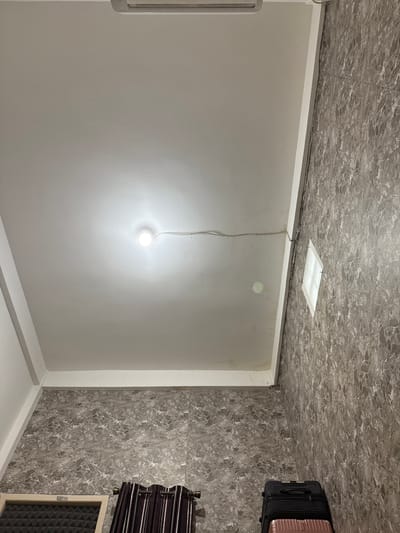 This image depicts an interior shot of a room, likely a bedroom or hotel room, with a view angled towards the ceiling and two adjacent walls. The ceiling is white and appears to be made of a smooth material, possibly gypsum board, with a single, bare light bulb illuminating the space. A thin electrical wire runs horizontally across the ceiling, attached at intervals. An air conditioning unit is visible at the top edge of the frame, partially obscured.
One wall is covered in a patterned wallpaper with a gray, marbled design. A small, square window with a translucent covering is set into this wall. The other wall, visible on the left, is also covered in the same marbled wallpaper.
In the lower portion of the image, on the right side, a stack of two suitcases is visible: a black hard-shell suitcase on top of a pink one. On the left side, a window frame with a textured acoustic panel behind it is partially visible, and next to it, a set of dark curtains with vertical stripes.
The overall impression is that of a functional, perhaps budget-friendly, lodging space. The image does not provide specific visual cues to pinpoint its exact location within Bandar Lampung, Indonesia, beyond being an indoor setting.