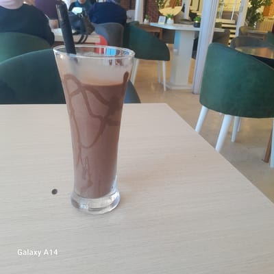 A tall glass of chocolate milkshake sits on a light-colored wooden table. The milkshake has a thick consistency with chocolate syrup drizzled down the inside of the glass. A black straw is partially submerged in the drink. The background shows a blurred scene of a cafe with green chairs and tables. The lighting suggests it's daytime. On the table, the text "Galaxy A14" is visible.