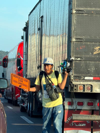 A man wearing a baseball cap, yellow t-shirt, and crossbody bag holds an orange sign written in Spanish "Memorias USB con 5000 canciones," meaning USB sticks with 5000 songs. He stands between lanes of traffic near a large truck. Other cars surround him, suggesting he's selling to drivers stuck in traffic. He is positioned in front of what appears to be an opened semi-truck trailer.