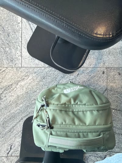 The image depicts an overhead shot of a gray tiled floor with a black leather seat on the left side and a green North Face backpack on the right. The backpack is partially obscured by a black object, possibly another seat or piece of equipment. The location appears to be indoors, potentially a gym or studio, but there are no indications of the city of Santa Cruz del Valle, Mexico.