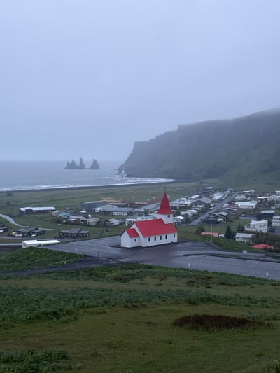 This image shows a rainy, overcast view of the village of Vik, Iceland, with the iconic Vik i Myrdal Church in the foreground. The church has white walls and a bright red roof. The village nestles between a steep, dark, green mountainside to the left and the Atlantic Ocean. In the ocean, there are several distinctive rock formations. The village comprises small houses and buildings, and a road leads along the hillside. Vegetation includes grass and low-lying plants.