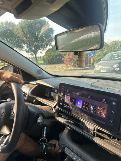 The image captures the interior of a car, looking forward through the windshield. The driver's hand is visible on the steering wheel. The car's dashboard features a large touchscreen display showing a car's infotainment system with various icons and text in Italian. Outside the car, trees and a road are visible, with another car in the distance. The scene appears to be a sunny day. The image was taken in Rudiano, Italy.