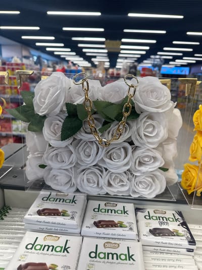 The image shows a display of merchandise inside what appears to be a retail store in Gaziosmanpaşa, Turkey. On the right, there are stacks of "Nestle damak" chocolate bars, including "Antep Fıstıklı Sütlü Çikolata" (Pistachio Milk Chocolate) and "gece" (night) variations. To the left of the chocolates is a clear, rectangular box filled with artificial white roses, adorned with green leaves and a gold chain handle. The roses are arranged densely within the box. The background suggests a store environment with rows of products and shelf lighting.