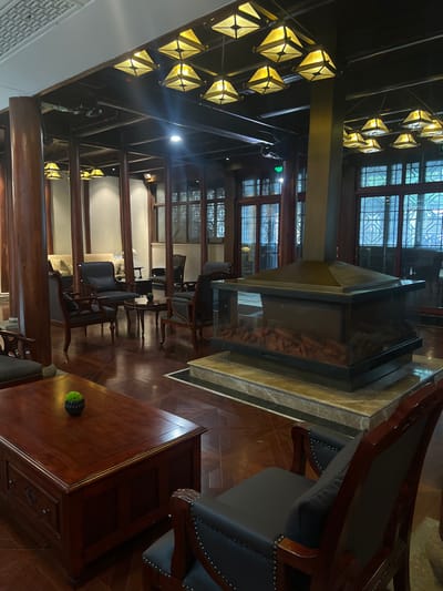 This image depicts an unoccupied interior lounge area, located in Tongxiang, China. The scene showcases a spacious room with dark wooden architectural elements, including numerous thick pillars and exposed ceiling beams. A central, square, four-sided glass electric fireplace with visible artificial embers is elevated on a multi-tiered light marble platform. A square, metallic chimney structure extends vertically from the fireplace to the ceiling.

Seating arrangements are distributed throughout the room, consisting primarily of dark wood armchairs with dark grey upholstered cushions and backs, often featuring decorative metallic studding. These chairs are positioned around dark wood coffee tables, some rectangular with drawers, and others round. In the background, lighter-colored upholstered sofa seating is visible against a wall.

The ceiling is equipped with multiple square-pyramidal light fixtures with yellowish glass panels, interspersed with recessed spotlights, providing illumination. Large, wooden-framed windows with intricate lattice designs are visible along the far walls, admitting ambient light. The floor is composed of dark wood parquet laid in a geometric pattern. A small green potted plant is centrally placed on a rectangular coffee table in the foreground. No individuals, actions, or interactions are present in the scene.