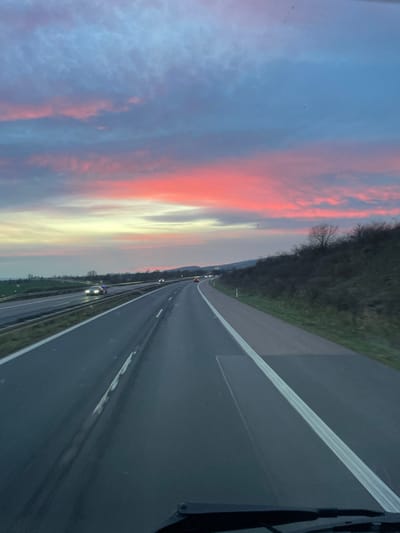 The image depicts a forward-facing view from inside a vehicle traveling on a multi-lane highway (Autobahn) during twilight in Wallhausen, Germany. The sky occupies the upper two-thirds of the frame, showcasing a vibrant display of deep blue, purple, pink, and orange hues with horizontal cloud formations, indicating either sunrise or sunset.

To the right of the highway, a dark, brush-covered embankment with sparse, leafless trees ascends. On the left, beyond the opposing carriageway, distant low-lying terrain is visible. The asphalt road surface appears damp, reflecting the ambient light. White solid lines delineate the shoulder, and dashed white lines separate the lanes; at least two lanes are visible in the direction of travel. The highway curves gently to the right in the mid-ground.

Several vehicles are present on the highway: multiple oncoming vehicles display illuminated headlights in the left lanes of the opposing carriageway, while faint red taillights of distant vehicles are visible in the lanes ahead of the observer. The bottom edge of the image reveals a portion of the observer's vehicle dashboard and a windshield wiper blade, confirming the perspective from within a moving vehicle. No individuals are explicitly discernible.