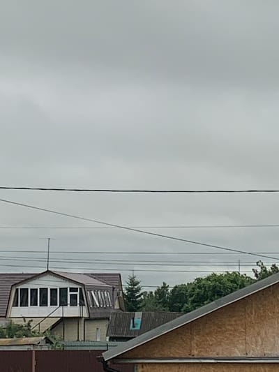 The image depicts a rural scene in Karmanovo, Russia, under an overcast sky, suggesting it's daytime with potentially poor weather. The primary subjects are buildings and power lines. On the right side of the frame, a portion of a house with an OSB-covered wall is visible, angled towards the viewer. Adjacent to it is another building with a dark tiled roof and a prominent section featuring multiple rectangular windows, some with white frames and dark panes, suggesting a living space. A smaller structure with a turquoise-colored window or vent is also visible on the roofline of this building.
Several power lines stretch horizontally across the image, dividing the sky from the buildings. In the background, lush green trees are visible, indicating a natural, possibly forested, environment surrounding the buildings. The overall impression is one of a quiet, possibly somewhat weathered, residential area. There are no people visible in the image. The time of day appears to be daytime, given the overall illumination, but the cloudy conditions obscure direct sunlight. No text is discernible within the image.