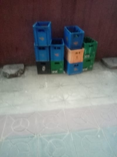 The image shows a stack of nine plastic crates of various colors (blue, green, orange, black) against a red wall. The crates are located on a tiled floor of alternating blue, white, and pink tiles with a pattern of circles inside squares. A rock or concrete fragment is visible to the left of the crates and another to the right. The photograph appears to have been taken indoors, although the specific location within Iyiowa Odekpa, Nigeria, is not discernible from the image. No people are present. The focus is somewhat blurry, obscuring finer details.