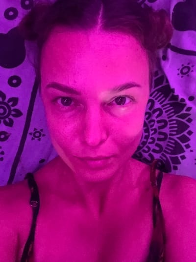 The image presents a close-up, high-angle portrait of a person, appearing to be a young adult female, lying down indoors. The scene is enveloped in striking magenta and purple artificial lighting, which saturates the person's skin and the surrounding environment with vibrant hues.

The subject's face, prominently featured, looks directly towards the viewer with a neutral to slightly contemplative expression. Dark eyebrows frame their eyes, which are clearly visible. Dark hair, possibly styled in buns or braids, is visible on the left side of the frame. A dark strap of clothing, likely from a tank top or camisole, is partially visible over the right shoulder.

Behind and around the person is a patterned fabric, which could be a bedsheet or tapestry. The pattern features intricate black designs, including mandala-like motifs, stylized stars, and circles, set against a background that appears purple under the artificial light.

The strong, colored lighting makes it impossible to determine the time of day, but it suggests an evening or nighttime setting. There are no overt location cues beyond the patterned fabric, which, given the context of Arambol, India, might suggest a bohemian or spiritually themed decor. No text is visible in the image.