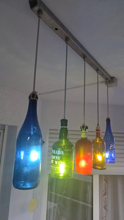 The image shows a close-up, low-angle shot of a homemade chandelier made from five glass bottles, each containing a light bulb. The bottles are suspended from a metal bar attached to a textured ceiling. From left to right, the bottles are dark blue, green, orange, amber, and bright blue. The green bottle has text that reads "STRADA LAWNY RT WINE". The orange bottle has text that reads "TRES P". The bright blue bottle has the word "Blue" printed on it. The lights inside the bottles cast colored glows. A yellow decorative item is visible hanging from the blue bottle on the far left. The background consists of window blinds and sheer curtains. The scene is indoors, likely a residence, and there are no people visible. The location is not specified in the image, and it is not possible to determine if it is in Crailsheim, Germany.