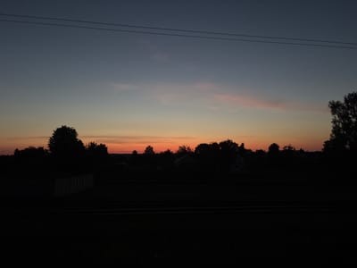 This is a photograph taken during twilight, likely near the village of Pratasevichi in the Pratasevicki Rural Council, Belarus. The sky transitions from a deep blue at the top to a gradient of orange and pink hues near the horizon, indicating the setting or rising sun. Two dark, thin power lines stretch horizontally across the upper portion of the image. The middle ground is dominated by a silhouetted landscape of trees, houses, and possibly a fence, all rendered in dark tones against the illuminated sky. In the foreground, two sets of parallel dark lines suggest railway tracks, with the area between and around them appearing as a dark, indistinct expanse, possibly fields or vegetation. There are no people or discernible actions visible in the scene.