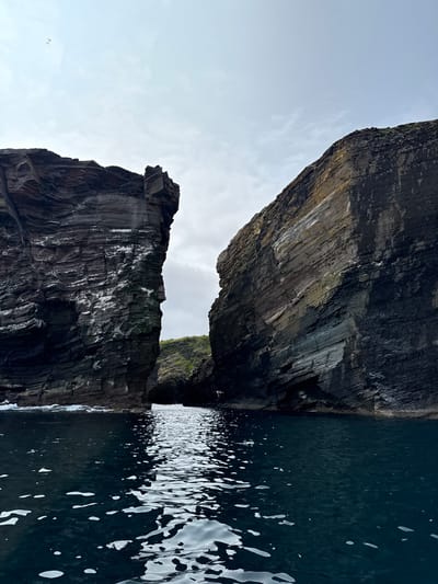 The image shows a view from the water looking towards a narrow sea passage between two imposing, dark, layered rock cliffs. The water in the foreground is dark blue and reflects the sky and cliffs. Small white waves are visible crashing against the base of the cliffs. The cliffs are rugged and textured, with visible horizontal striations. Patches of white, likely bird droppings, are scattered across the cliff faces. Green vegetation can be seen at the top of the cliffs in the distance. The sky is overcast with some clouds. Two birds are visible flying in the sky in the upper left portion of the image. This scene is characteristic of the dramatic coastal geology found in Shetland, United Kingdom, likely depicting a cliff face or sea stack formation common in the region.