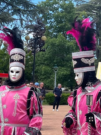 Two people dressed in elaborate Venetian costumes stand in a park-like setting in Saint-Galmier, France. The costumes are predominantly pink and black, heavily adorned with silver trim, jewels, and ornate headpieces with black and pink feathers. Both figures wear white masks. In the background, other people can be seen walking on a paved path, along with trees and street lamps. The scene is captured outdoors during the daytime.