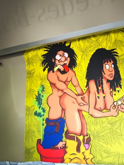 The media file displays a cartoon illustration on a textured yellow fabric, likely a tapestry or wall hanging, set against a neutral, textured background with a silver strip running horizontally across the top. The illustration features two cartoon characters, one male and one female, with dark dreadlocks and attire inspired by Rastafarian culture. The male character has brown skin, thick eyebrows, and his tongue is sticking out. He is wearing blue overalls, partially pulled down, and stands in a red pot, from which a stylized green plant grows. The female character also has brown skin and dreadlocks adorned with beads in red, yellow, and green. She is depicted from the waist up, leaning forward, with her hands held out. The background of the illustration is a vibrant yellow with a repeating pattern of cannabis leaves. The scene appears to be set in an indoor location, given the overhead lighting and the nature of the fabric display. There is no visible text within the image. The overall mood of the illustration is playful and whimsical.