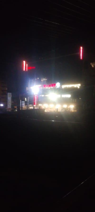 The image shows a nighttime view of a building with illuminated signage. The prominent sign reads "K MART" in red letters, with a white floral or star-like symbol to its right. Several other illuminated signs and lights are visible on the building and in the surrounding area, suggesting it is a commercial establishment. Red lights are also visible on poles or structures flanking the building. The foreground is dark, indicating the photo was taken from a distance or in a dimly lit area. The scene appears to be located in an urban or commercial district.