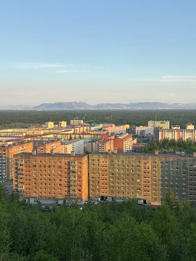This is a vertical, eye-level, medium-range shot of an urban landscape featuring rows of apartment buildings and dense green trees. The scene is bathed in the warm, soft light of either sunrise or sunset, casting long shadows and highlighting the textures of the buildings. The sky is a pale blue, fading to a lighter hue near the horizon.
The primary subjects are the numerous apartment blocks, which vary in color and design. Some are a muted green, while others are painted in shades of yellow, orange, and pale pink, some appearing to be under construction or renovation. The buildings are tall and densely packed, interspersed with a thick band of green trees that form a natural barrier between the residential areas and a distant, hazy mountainous landscape. The trees are lush and full, suggesting a temperate climate during warmer months.
The activity appears to be a quiet, static view of the city. There are a few vehicles visible on the road between the buildings, and some individuals can be seen on the sidewalks or near building entrances, but they are too small to discern details. The overall atmosphere is serene and somewhat stark, reflecting the imposing architecture and the vastness of the surrounding natural environment.
Notable details include the consistent grid-like arrangement of windows on the apartment buildings, many of which appear to be the same design. The lighting suggests a time of day when the sun is low, possibly early morning or late evening, creating a dramatic contrast between light and shadow. The presence of mountains in the distance and the type of vegetation hint at a location in a northern latitude with distinct seasons. No visible text is present in the image. This scene is consistent with the urban environment of Norilsk, Russia, known for its stark architecture and surrounding natural beauty.