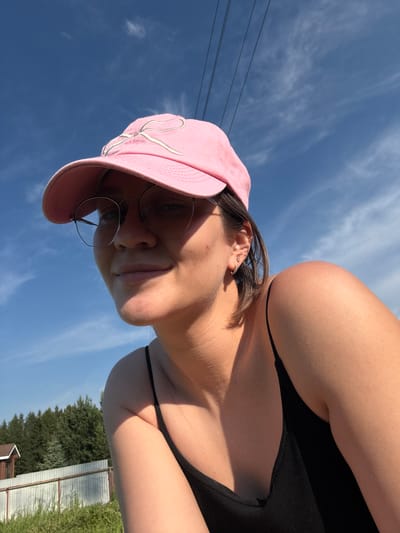 In a low-angle, outdoor selfie, a young woman wearing a pink baseball cap and round wire-frame glasses is pictured against a bright blue sky with scattered clouds and visible power lines. She is looking towards the camera with a slight smile, her head tilted. She has short brown hair and is wearing a black tank top. The background shows a blurred natural setting with trees, a fence, and a small building, suggesting a rural or suburban environment. The lighting indicates it is daytime, likely sunny.