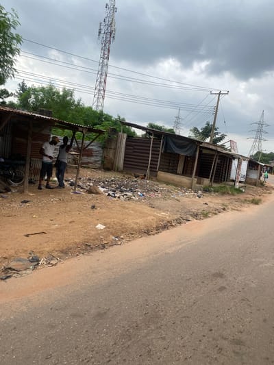 In Ojere, Nigeria, a dirt road runs alongside a row of makeshift shops. The shops have corrugated metal roofs and walls, with some open fronts revealing their interiors. In front of the shops, there is a significant amount of litter, including plastic bags, scattered debris, and what appears to be household waste. Two men are standing under the roof of one of the shops, appearing to converse. A motorcycle is parked to the left of the shops. In the background, a tall communication tower and several electricity pylons are visible against a cloudy sky. The environment appears somewhat neglected, with a general atmosphere of disarray due to the accumulated refuse.