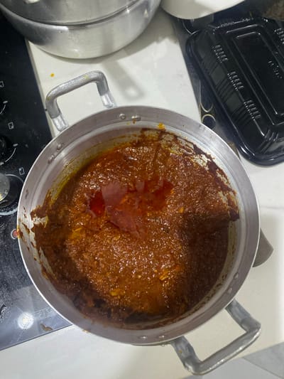 The image displays an overhead view of a large, silver-colored metal pot filled with a thick, dark reddish-brown sauce or stew. The sauce has an oily sheen on its surface, suggesting it is either actively cooking or freshly prepared. Small, lighter-colored chunks, likely pieces of ingredients such as meat, fish, or vegetables, are visible within the rich mixture. The pot is equipped with sturdy, U-shaped metal handles on its sides.
The cooking vessel is placed on a white countertop or stovetop. A black electric or gas hob, with visible control knobs, occupies the lower part of the frame. To the upper left of the main pot, a black plastic takeaway container or its lid is partially visible. Another silver metal pot or lid is partially seen on the far left, further indicating a kitchen setting. The appearance of the dish is consistent with traditional West African cuisine, such as a hearty stew or soup, aligning with the provided location context of Adenta Municipality, Ghana. No people are present in the image, and there is no visible text. The time of day and weather cannot be determined.