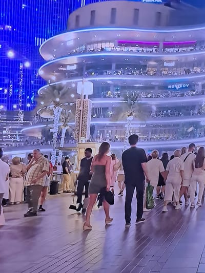 The image depicts a bustling urban scene, likely at night given the vibrant artificial lighting. In the background, a multi-tiered, circular building with illuminated windows and balconies is visible. The architecture is modern, with sweeping curves and glowing accents. Palm trees with decorative lights are interspersed along the ground level, suggesting a warm climate or an indoor botanical display within a large structure.
In the foreground, a crowd of people are walking on a tiled pathway. The individuals are dressed in casual attire. Several couples are seen walking together, holding hands. One woman in a green dress is walking away from the camera, while a man in a black shirt and pants walks beside her. Other people are seen in various groupings, some walking towards the camera, others moving away. The overall atmosphere appears to be one of a public gathering, perhaps a promenade or a marketplace.
There is no indication that this image is located in Cēsis, Latvia. The architecture and vegetation do not align with the known characteristics of Cēsis.