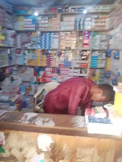 The image displays the interior of a retail establishment located in Panshekara, Nigeria. A male individual, wearing a maroon long-sleeved garment, stands behind a wooden counter, leaning forward and looking down at items placed on its surface. The counter, which occupies the lower foreground, exhibits wear and has scattered white markings. Behind the individual, a white plastic chair with a beige backrest is partially visible. The background consists of multiple shelves packed densely with a variety of packaged goods, including what appear to be pharmaceutical products, toiletries, and stacks of notebooks or paper. An overhead light fixture illuminates the cluttered shelving. On the counter in front of the individual, several boxes and packages, including white, blue, and yellow items, are stacked.