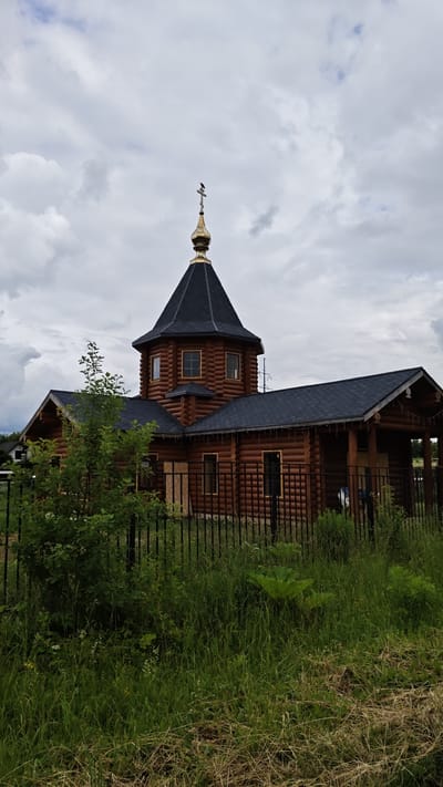 This image depicts a wooden Orthodox church in Sof'ino, Russia. The church features a prominent, dark-shingled roof with a golden dome topped by a cross. A bird is perched on the cross. The walls of the church are constructed from rounded logs, giving it a rustic appearance. Several windows with golden trim are visible, and along the eaves of the main structure, there are decorative string lights.

The church is set against a backdrop of a cloudy, overcast sky, suggesting either early morning or late afternoon, or simply a day with diffused light. In the foreground, a black metal fence encloses the church grounds, with lush green grass and various plants growing tall and wild behind it. A leafy green bush is situated to the left of the church, partially obscuring the view of its side. The overall scene conveys a sense of tranquility and perhaps a touch of solemnity due to the overcast weather. There is no visible text in the image.