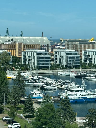 The image shows a harbor in Gdynia, Poland, with numerous boats and yachts docked. In the background, modern buildings and industrial structures line the waterfront. A large blue and white ship is prominently featured in the foreground. The sky is clear and blue, indicating a sunny day. The scene appears to be a bustling port with a mix of leisure and commercial activities.