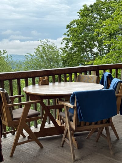 The image shows an outdoor dining area on a porch or balcony with a wooden table and chairs. Blue blankets are draped over some of the chairs. A condiment set rests on the table. The background features a green landscape with trees and a view of mountains under a partly cloudy sky. The location of the image in the city of Progress, Russia is not supported by the visual content.