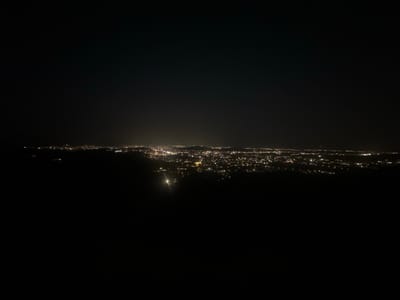The image shows a panoramic view of a city at night, likely from an elevated vantage point. The city lights are spread across the horizon, creating a tapestry of glowing dots against the dark sky. The foreground is dominated by the darkness of what appears to be a hilly or mountainous terrain, with only the faintest outlines visible. One distinct light source, possibly a streetlamp or a vehicle's headlights, is visible in the mid-ground, illuminating a small patch of the dark landscape. The overall atmosphere is one of quiet and expanse, with the city lights representing the bustling life within the vast darkness. Without additional information, pinpointing the exact location within Grbavci, Montenegro, is not possible based solely on the visual data.