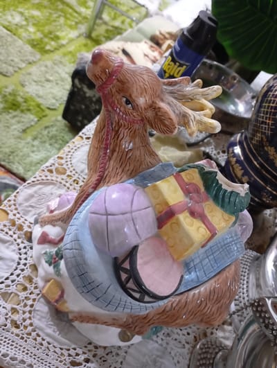 The scene depicts an indoor setting in Nasiriyah, Iraq. A brown ceramic reindeer figurine, featuring light brown antlers and a red harness, is centrally positioned on a white crocheted lace tablecloth. The reindeer is depicted carrying or pulling a light blue bundle filled with miniature objects, including a light pink basketball, yellow rectangular gift boxes with red ribbons, and a pink drum with black trim. To the right of the figurine, a dark blue decorative object with gold patterns and a reflective silver item are partially visible. In the background, a blue aerosol can labeled "Raid" stands upright next to a dark green artificial leaf. A light green patterned rug or carpet is visible further in the background.