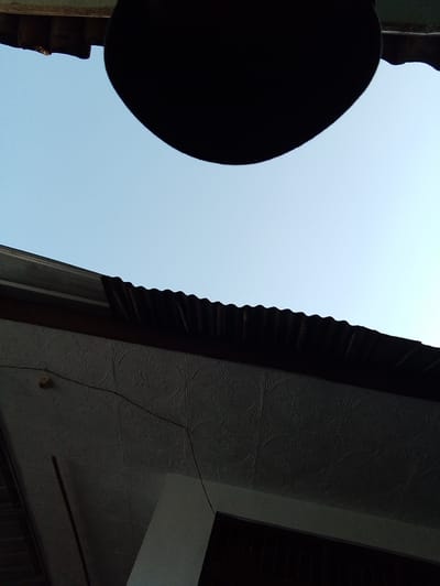 This is an exterior, ground-level, low-angle shot of a portion of a building's roof and the sky above. The sky is a clear, pale blue. A large, dark, circular object, possibly a hat or a piece of fabric, is in the upper portion of the frame, obscuring a portion of the sky. Below the sky is a corrugated metal roof that appears weathered. Beneath the corrugated roof is a textured ceiling, possibly made of stucco or a similar material, with a repeating floral or circular pattern. A thin wire runs diagonally across the textured ceiling, attached to a small, brown, possibly electrical, fitting. A thin, dark, vertical object, likely a pole or a stick, leans against the textured ceiling on the left side of the frame. The building appears to be located in Sukamanah, Indonesia. The scene is static and shows no people or activity.
