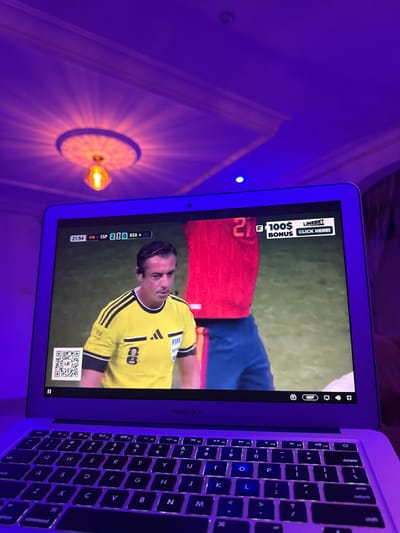 The image shows a laptop displaying a soccer match. The laptop screen shows a scoreboard with "ESP 2" and "RSA 0" indicating the score. There is a referee in a yellow jersey with the FIFA logo, and another player in a red jersey with the number 21. An advertisement for "LINEBET BONUS CLICK HERE!" is visible on the right side of the screen. A QR code is also present on the left side. The laptop is open and placed on a surface, likely a table, with a keyboard visible in the foreground. The scene is illuminated by a purple light, casting shadows on the ceiling, which has decorative molding. The overall setting suggests an indoor environment, possibly a living room or a dedicated viewing area. The specific location of Laminga, Nigeria, is not evident from the visual information in the image.