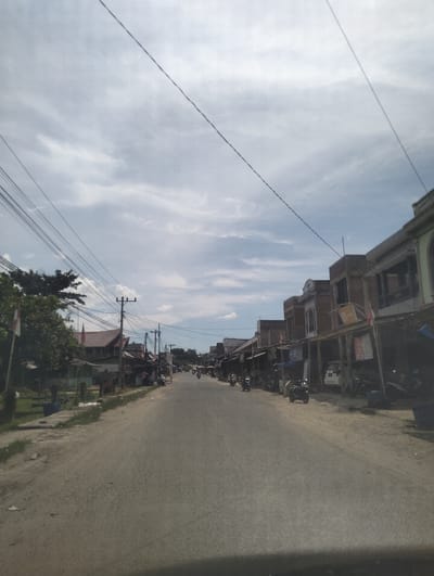 The image shows a straight, paved road lined with small buildings on the right side and trees and more buildings on the left. The sky is partly cloudy. Several utility poles with numerous wires are visible throughout the scene. On the right side of the road, there are shops with covered frontages, some displaying signs and advertisements. A white car is parked in front of one of the buildings, and several motorcycles are parked or being ridden on the road. On the left side of the road, there is a large tree with a flag attached to it, and more buildings and vegetation. The road appears to be a main thoroughfare in a semi-urban or rural setting. The overall impression is that of a typical day in a bustling street in Pulorayeuk, Indonesia.
